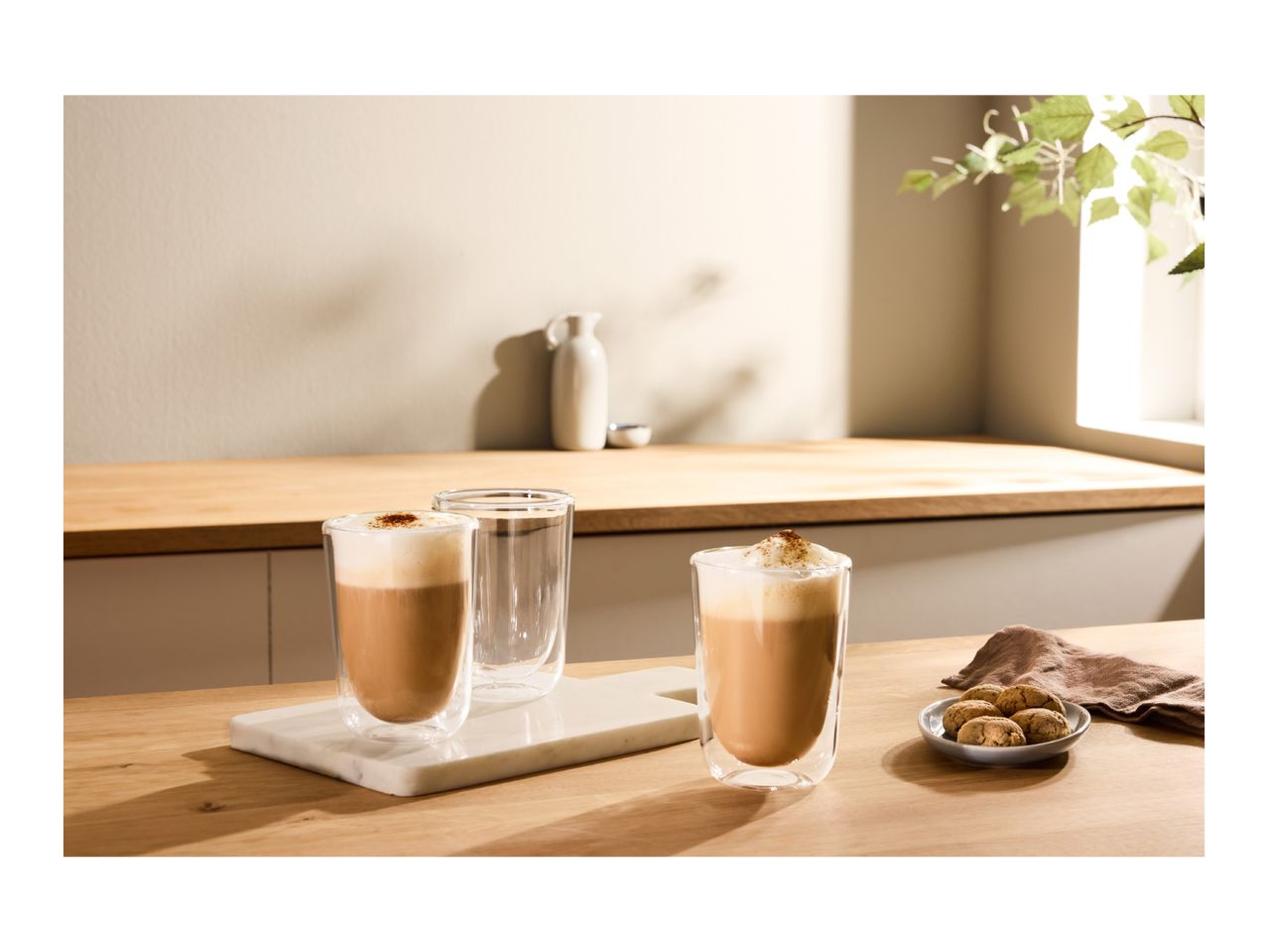 Two glasses of coffee with foam and a plate of cookies on a wooden counter.