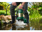A person in waders places a green submersible pump into a pond, causing water to splash.