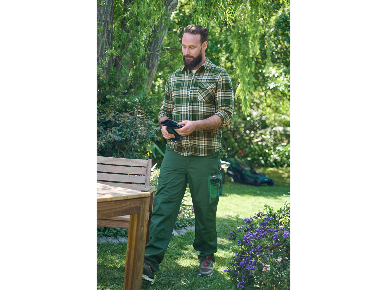 Man in work trousers and shirt examines gardening gloves in garden.