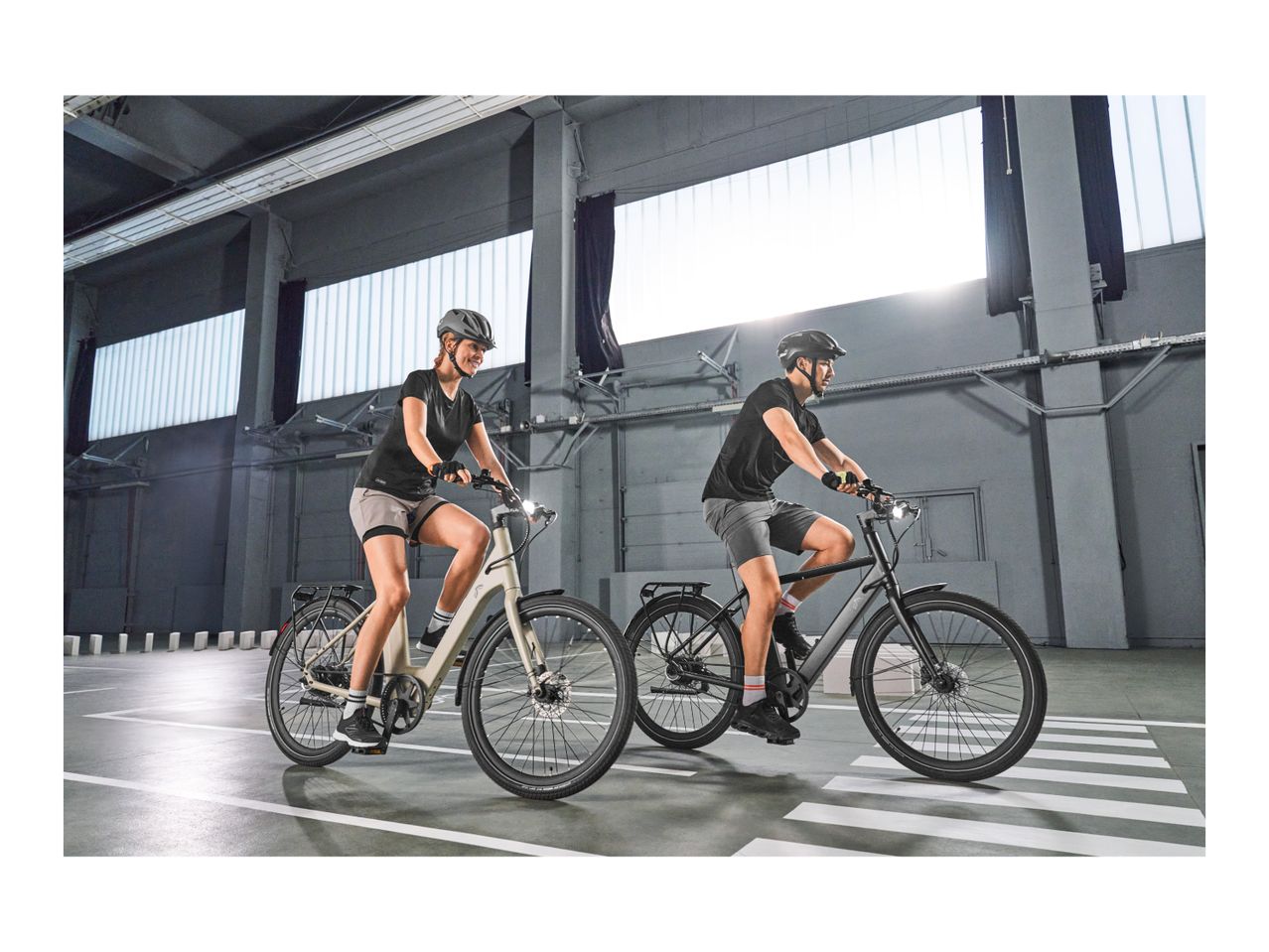 Two people riding electric bikes in an indoor parking garage with bright windows.