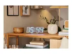 Digital clock displaying 15:44 on a wooden bedside table with a plant, books, and a lamp.