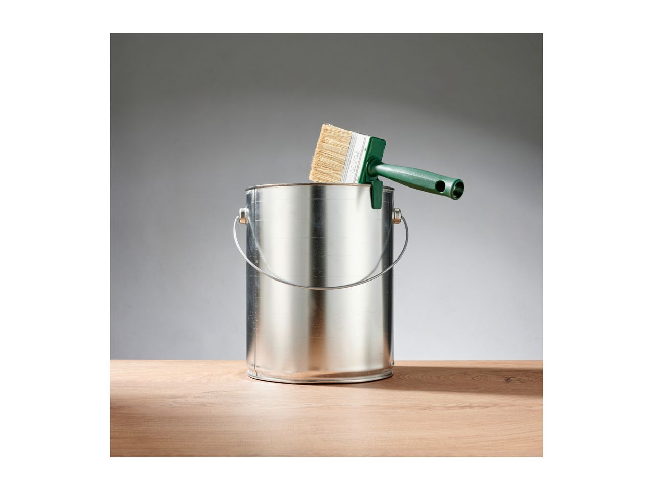 Paint can with a green-handled paintbrush resting on its rim, on a wooden surface.