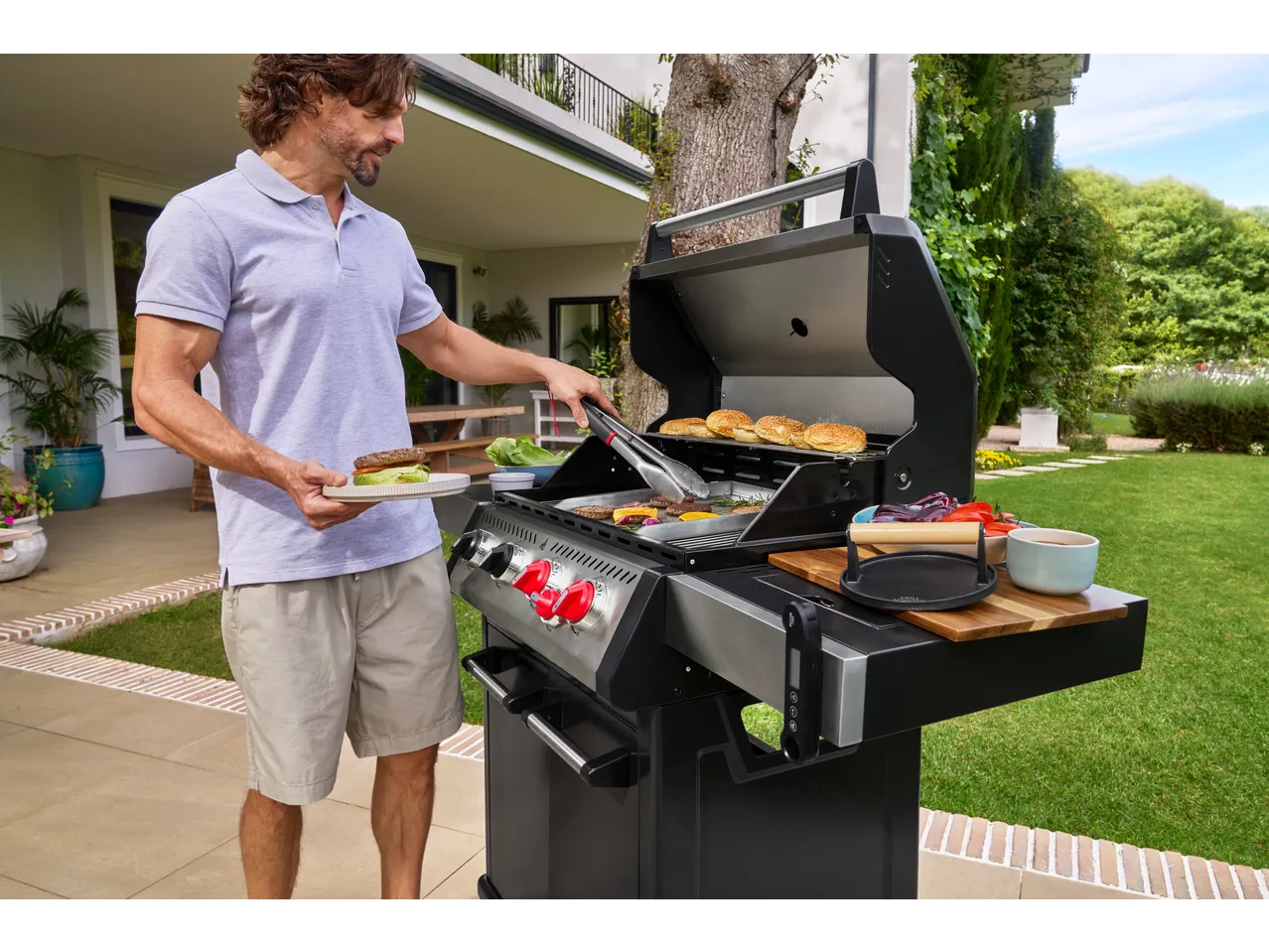 Bărbat gătind burgeri și chifle pe un grătar cu gaz în grădină.