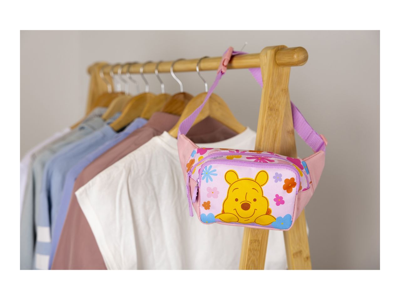 Pink fanny pack with a cartoon bear and flowers hanging on a wooden clothes rack with various shirts.