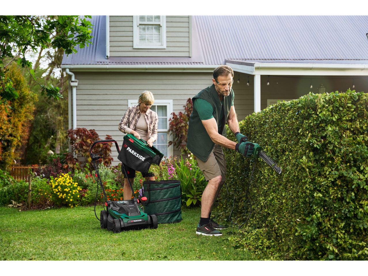 Man trimming hedge, woman emptying Parkside® Elektro-Rasenmäher lawnmower bag.
