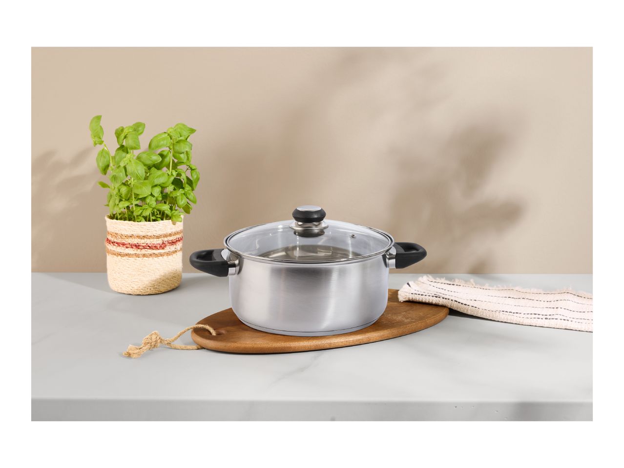 Stainless steel pot with a glass lid on a wooden board, next to a basil plant.
