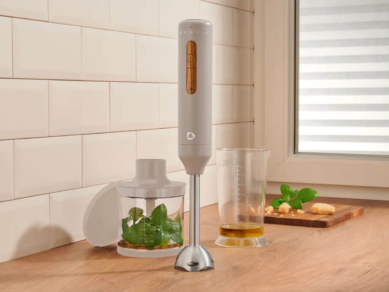 Hand blender with accessories, basil, parmesan, and olive oil on a kitchen counter.