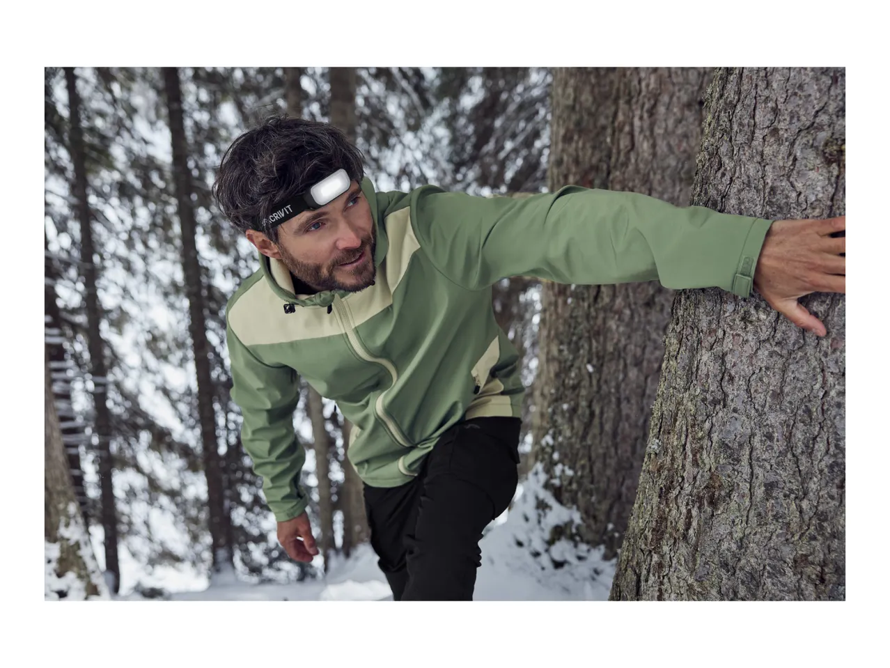 A man wearing a head torch and a green outdoor jacket in a snowy forest.