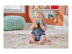 A baby in a denim romper sits on a patterned rug, chewing on a colorful teether toy.