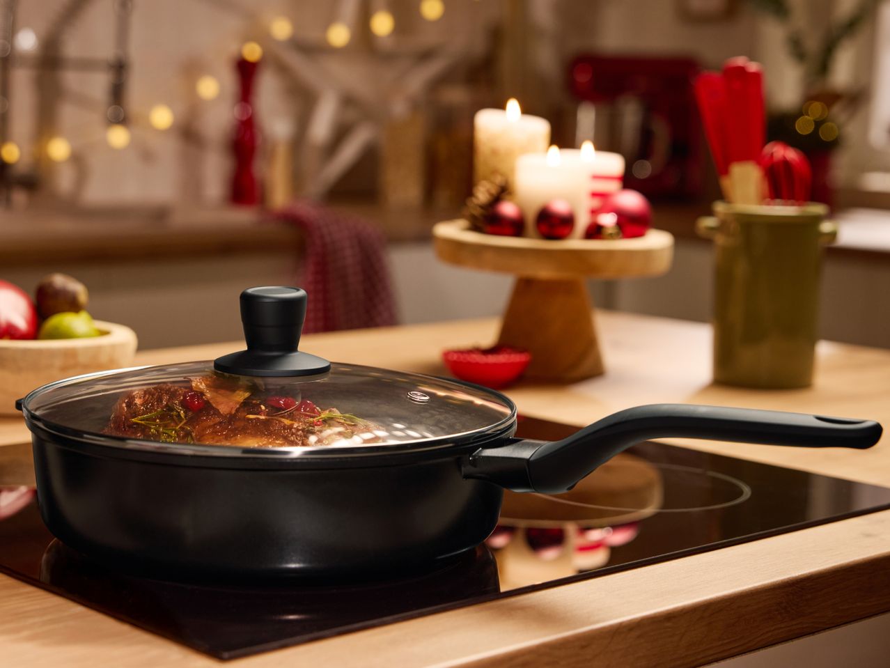 Black cooking pot with glass lid on an induction hob, with Christmas decorations in the background.