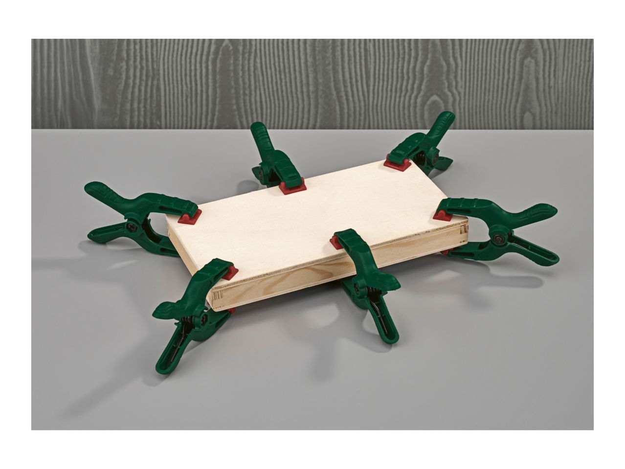 Six spring clamps holding together two pieces of wood on a grey surface.