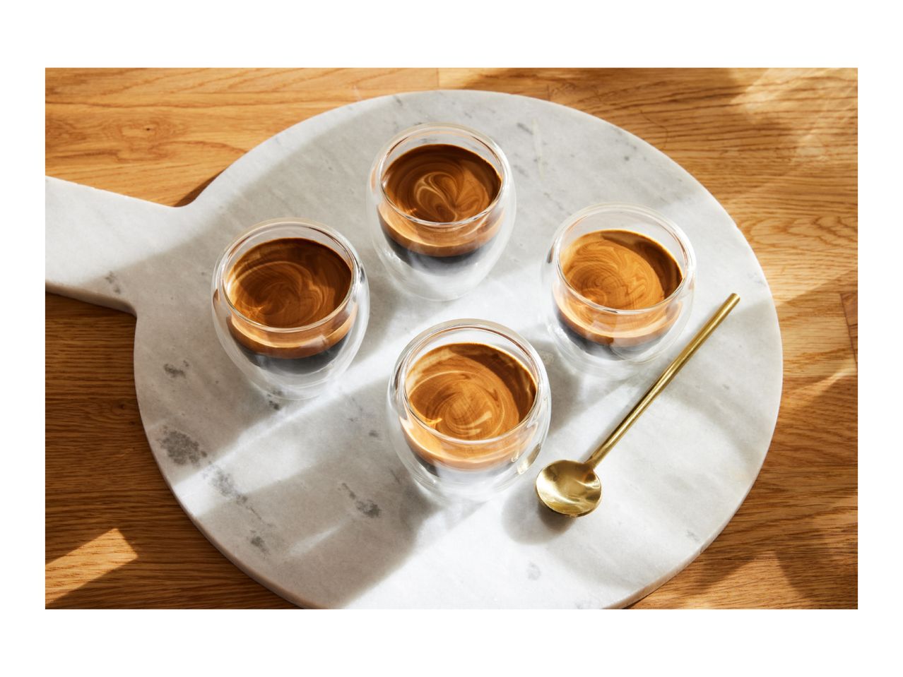 Four espresso shots in clear glasses on a marble serving board with a gold spoon.