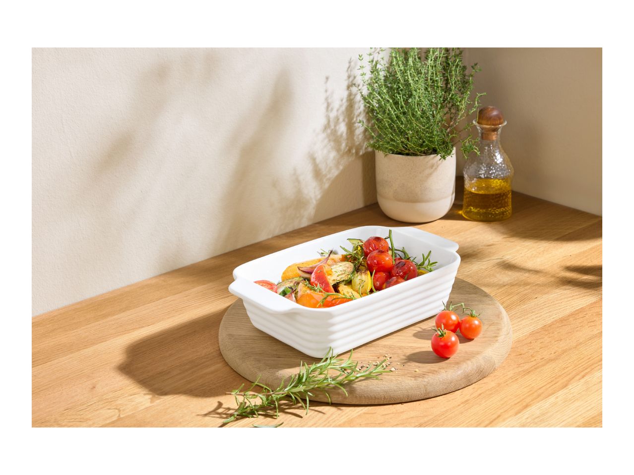 Roasted vegetables in a white baking dish on a wooden board, with herbs and olive oil.