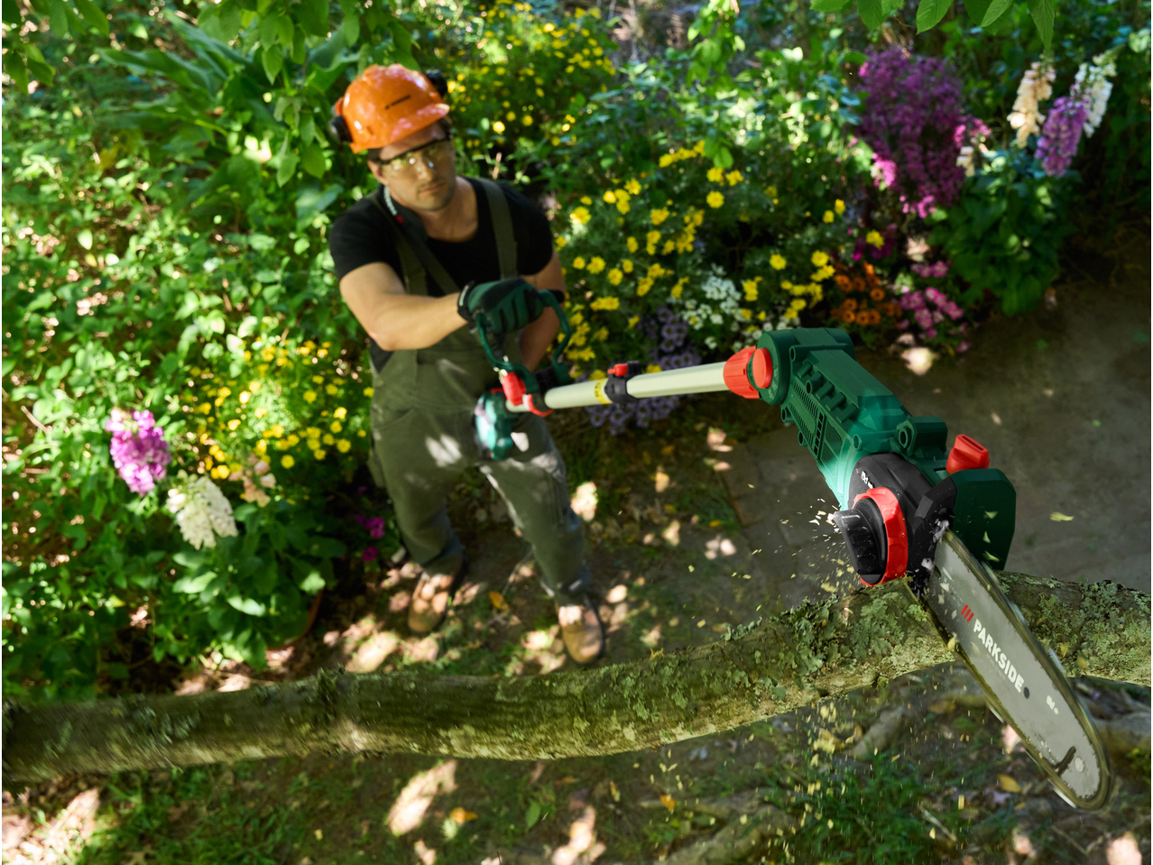 Man in safety gear using a pole chainsaw to cut a tree branch in a garden.