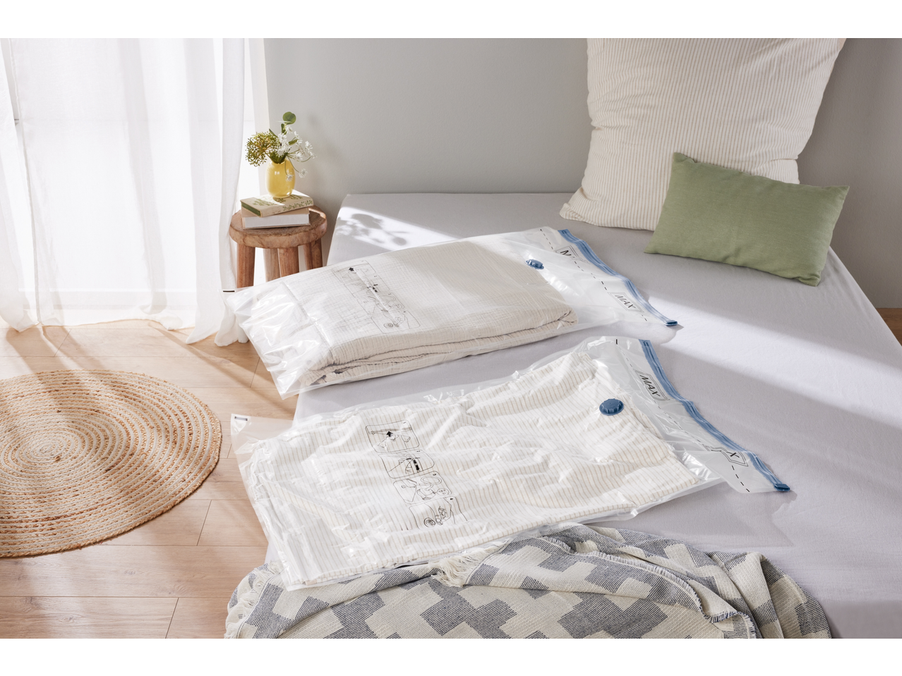 Two vacuum storage bags with textiles on a bed, next to a stool with a vase and books.