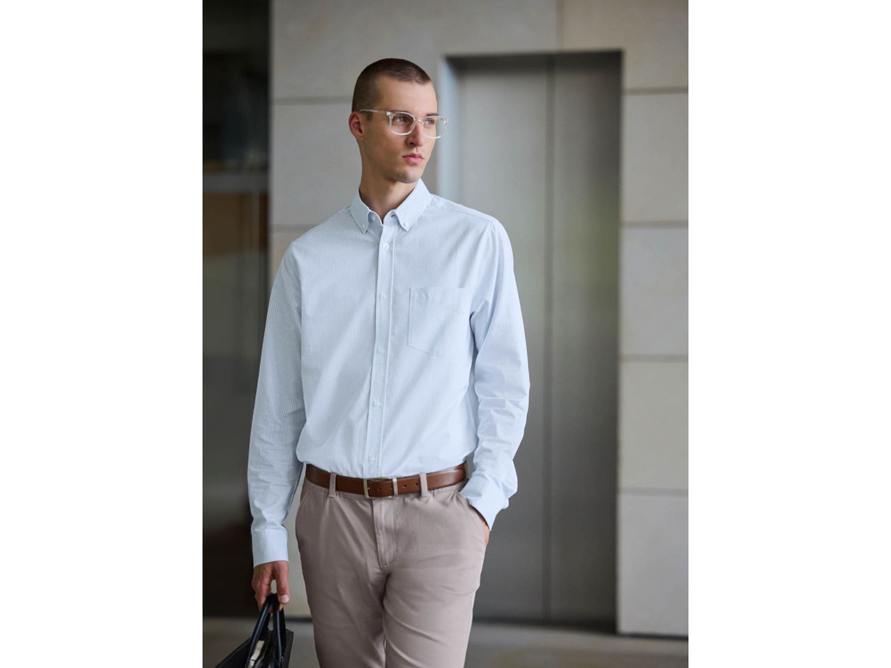 Man in light blue striped shirt, tan pants, and clear glasses, holding a bag.