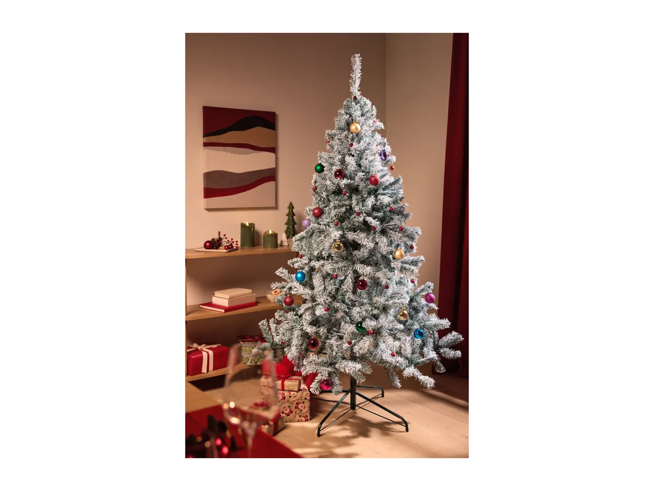 Flocked Christmas tree with colorful baubles, gifts, and festive decorations in a living room.