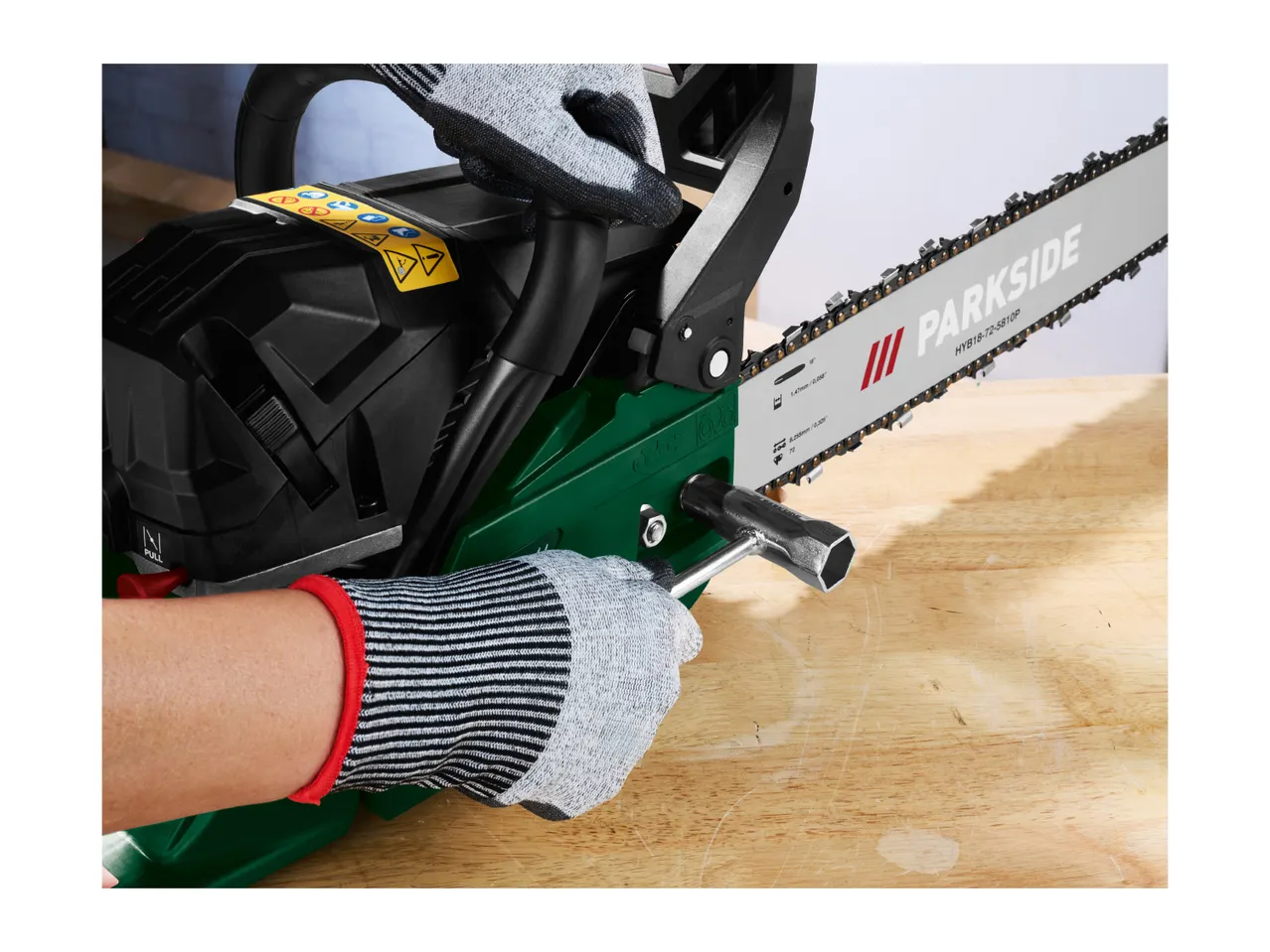 A person wearing protective gloves uses a wrench to adjust a chainsaw on a wooden table.