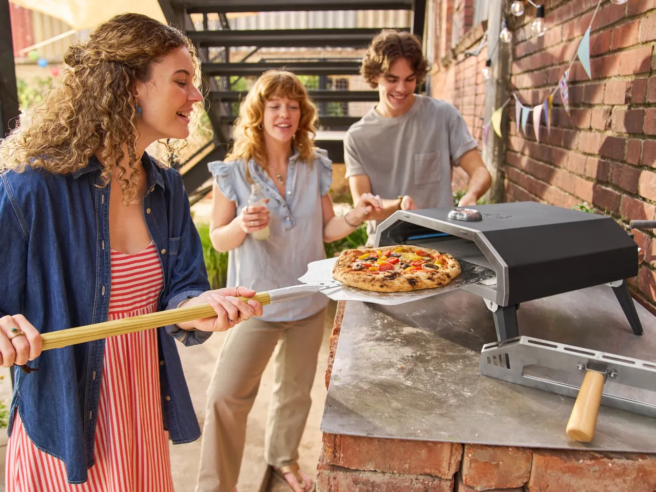 Tre amici preparano una pizza con un forno per pizza da esterno, su un tavolo di mattoni.
