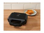 Black waffle maker on a wooden counter with two waffles on a plate in the background.