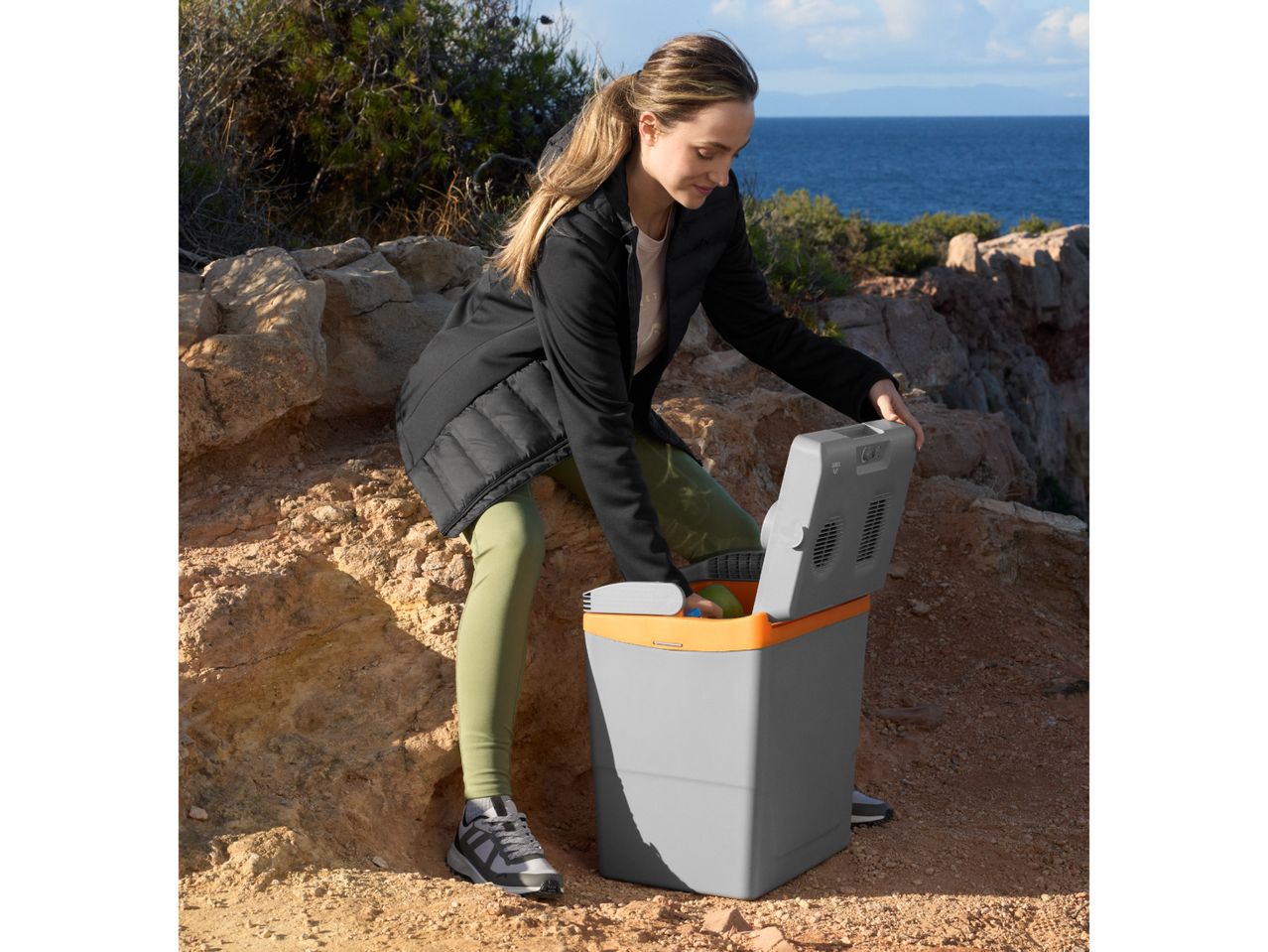 Woman opening a portable cooler on a rocky coastal path with the sea in the background.