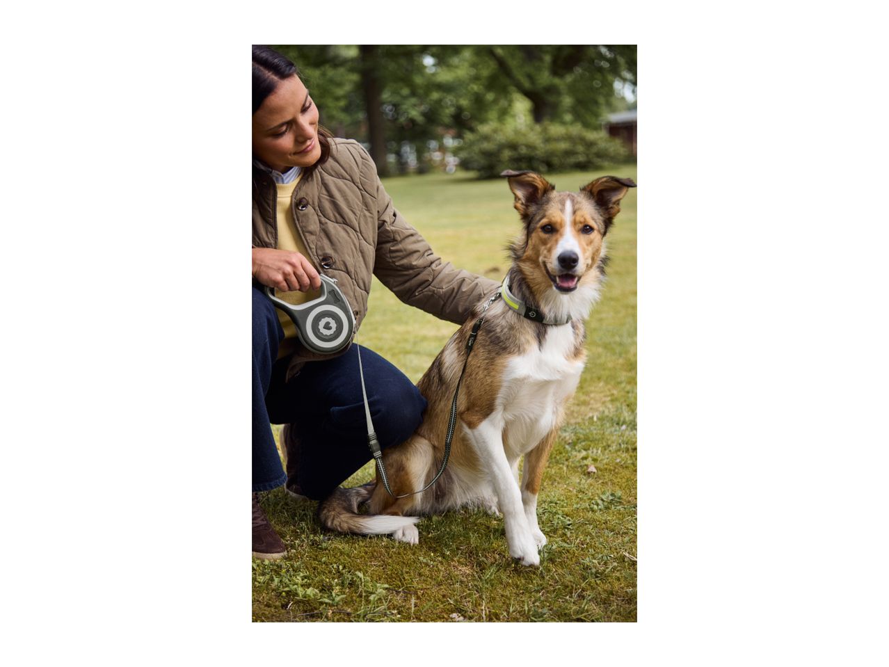 A woman holding a retractable dog leash next to her dog sitting on the grass.