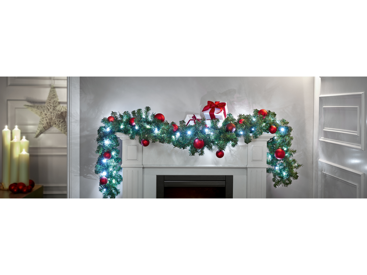 Christmas garland with red baubles and white lights draped over a white fireplace.