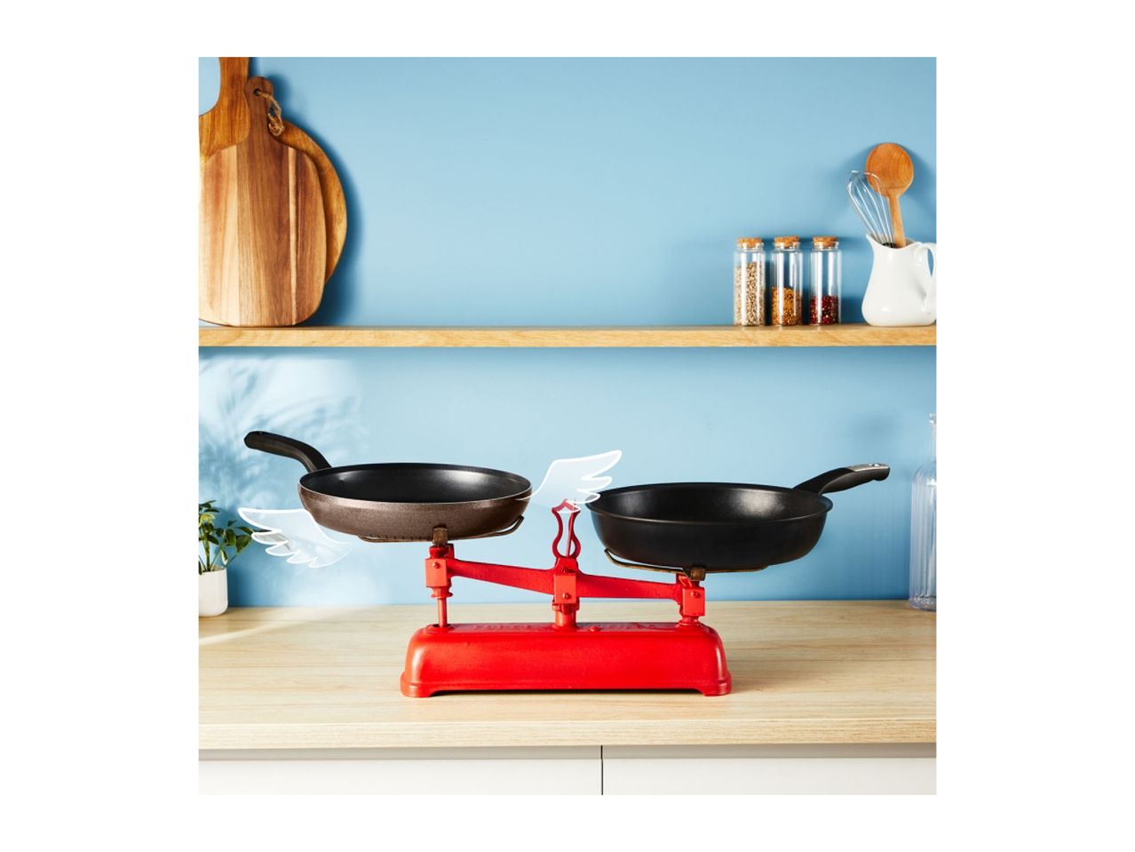 Red kitchen scale with two frying pans, on a wooden counter with kitchen utensils.