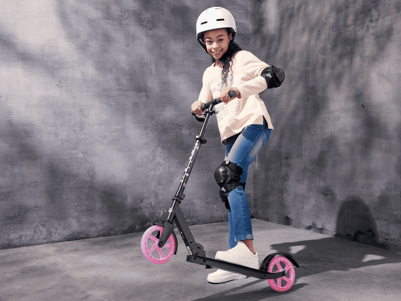Ein Mädchen fährt auf einem schwarzen Scooter mit rosa Rädern und trägt einen Helm und Schützer.