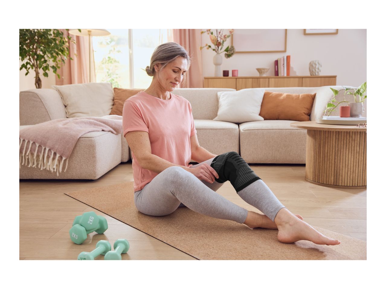 Woman wearing a knee support, sitting on a yoga mat with dumbbells nearby in a living room.