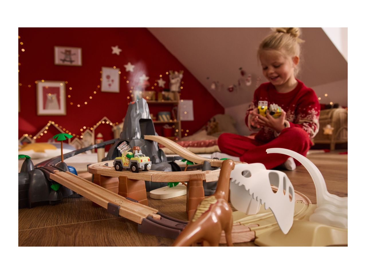 A child plays with a dinosaur-themed wooden train set and toy cars in a festive room.