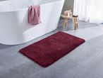 Un tappeto da bagno bordeaux su un pavimento piastrellato grigio, con una vasca da bagno bianca e uno sgabello in legno.
