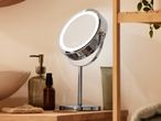 Illuminated makeup mirror with skincare products and a plant on a wooden shelf.