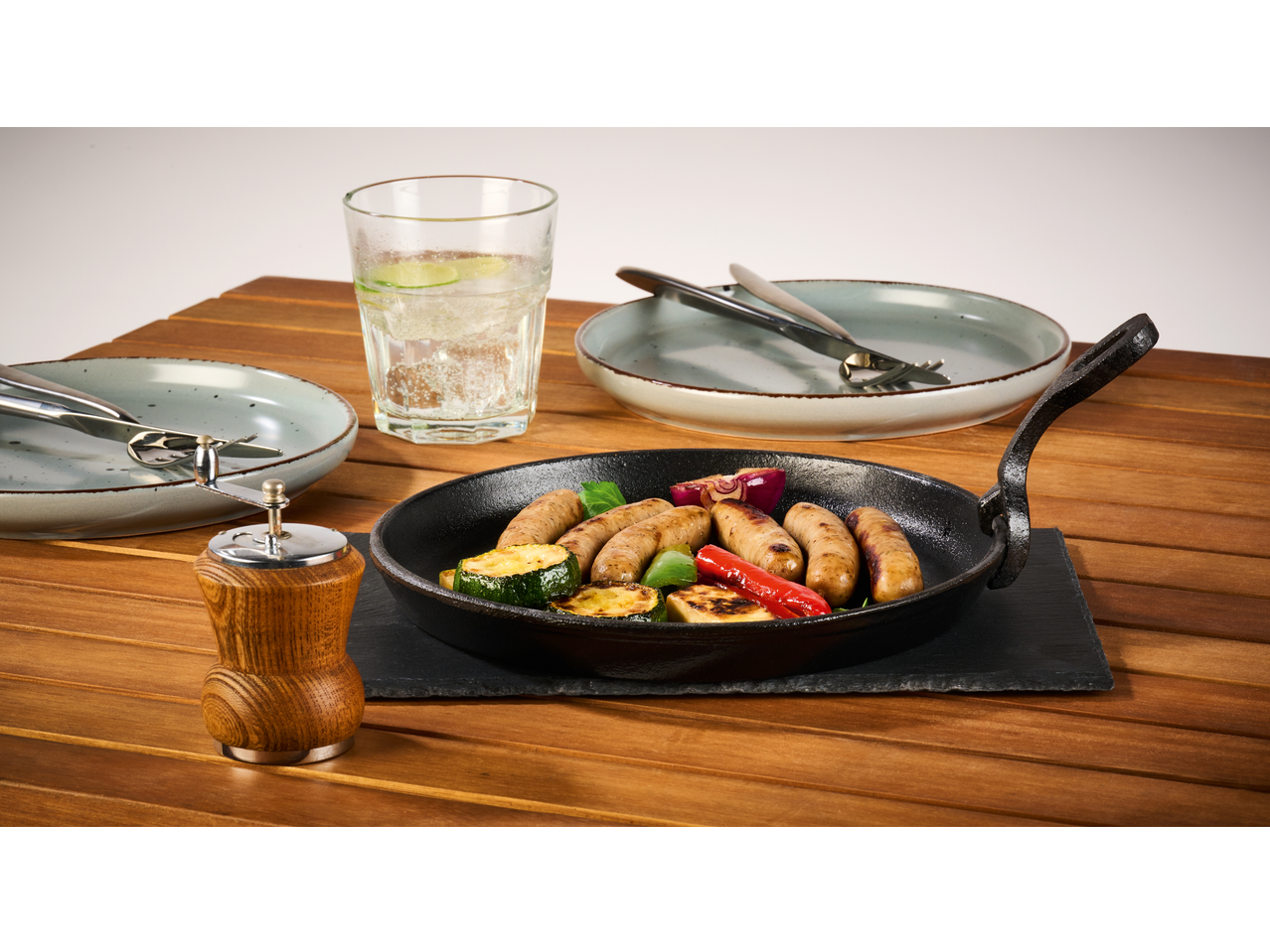 Tigaie din fontă cu cârnați și legume, un pahar cu apă de lămâie și farfurii pe o masă de lemn.