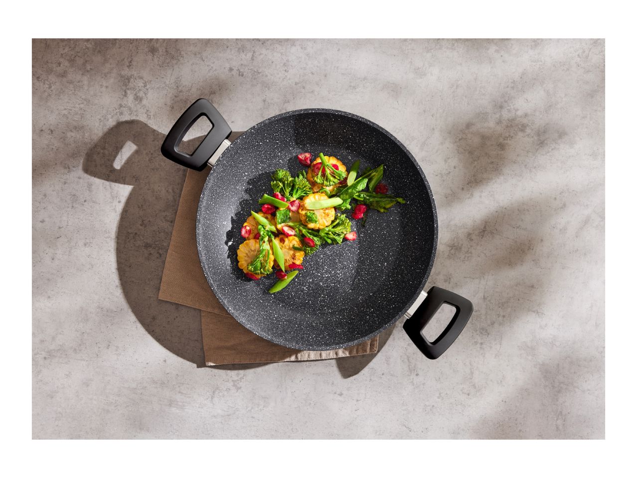 Overhead shot of a black granite wok with vegetables on a concrete surface.
