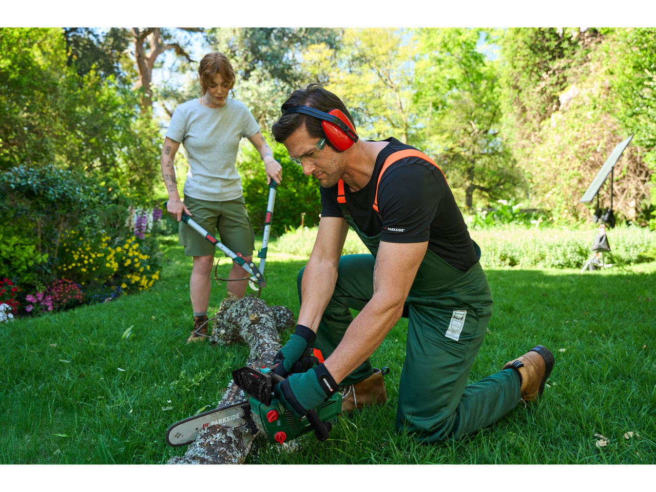 Moški reže deblo z motorno žago, ženska obrezuje z vrtnimi škarjami na vrtu.