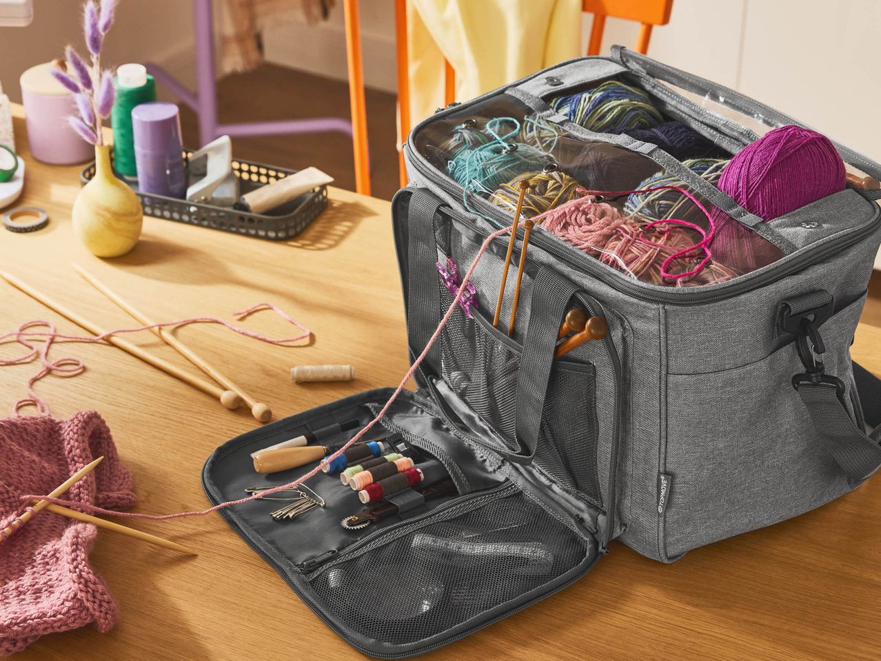 A gray yarn storage bag filled with colorful yarn and knitting supplies on a wooden table.