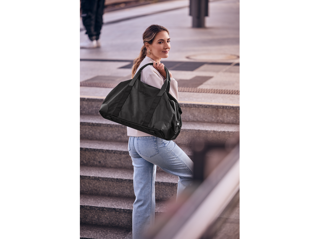Woman in jeans and white top carrying a black Esmara Weekend Bag on stairs.