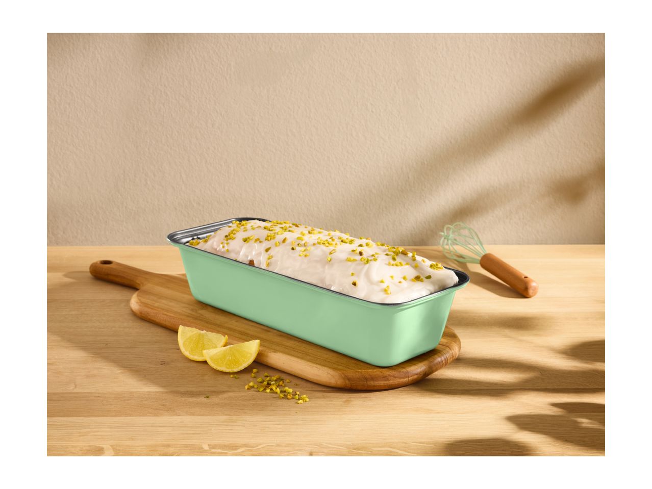 Lemon loaf cake with pistachio topping in a green baking pan on a wooden board.