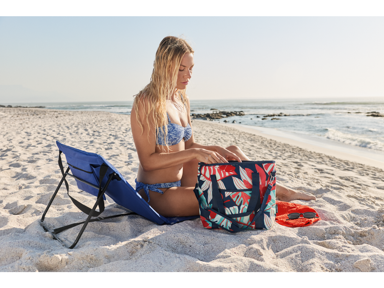 Woman in bikini on beach with CRIVIT cool bag and blue beach chair.