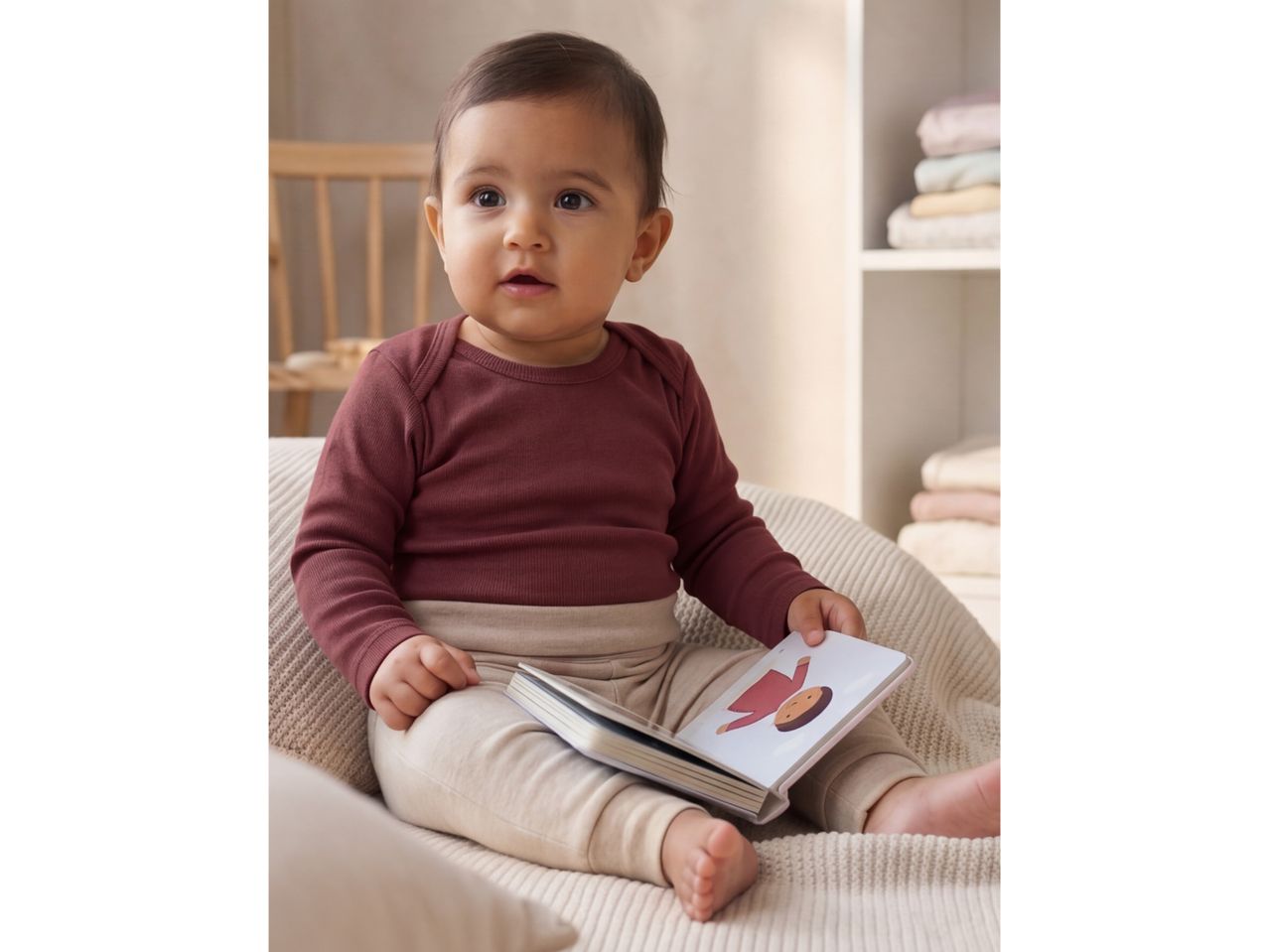 Baby in a long-sleeved bodysuit and leggings, holding a board book with an illustration of a person.