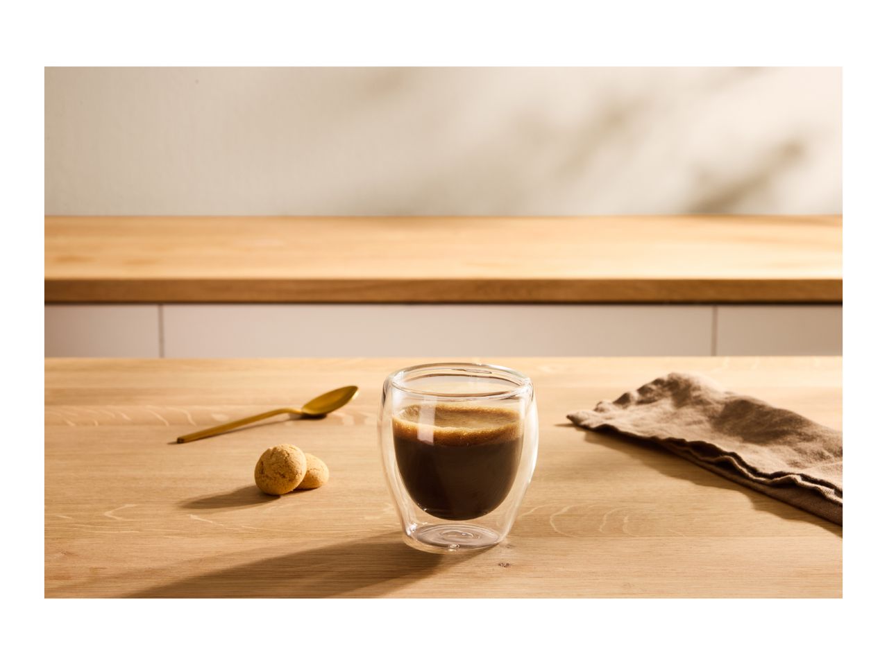 Espresso in a double-walled glass, with amaretti biscuits and a golden spoon on a wooden table.