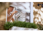 Mulher regando plantas num vaso branco com um regador de metal, com uma rede mosquiteira visível.