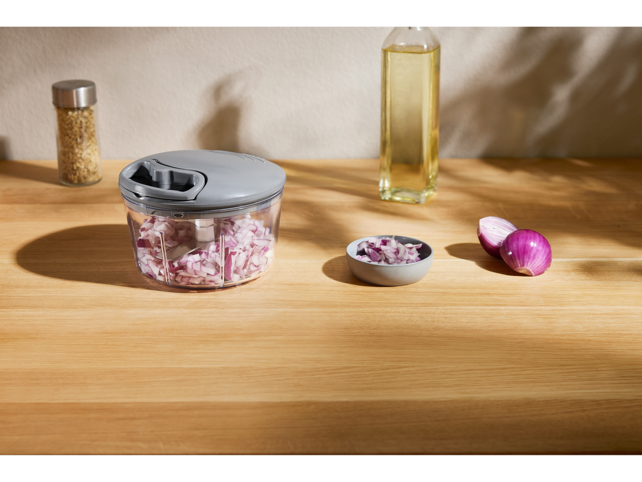 Ръчен кухненски чопър с нарязан червен лук; малка купичка и бутилка олио на дървена повърхност.