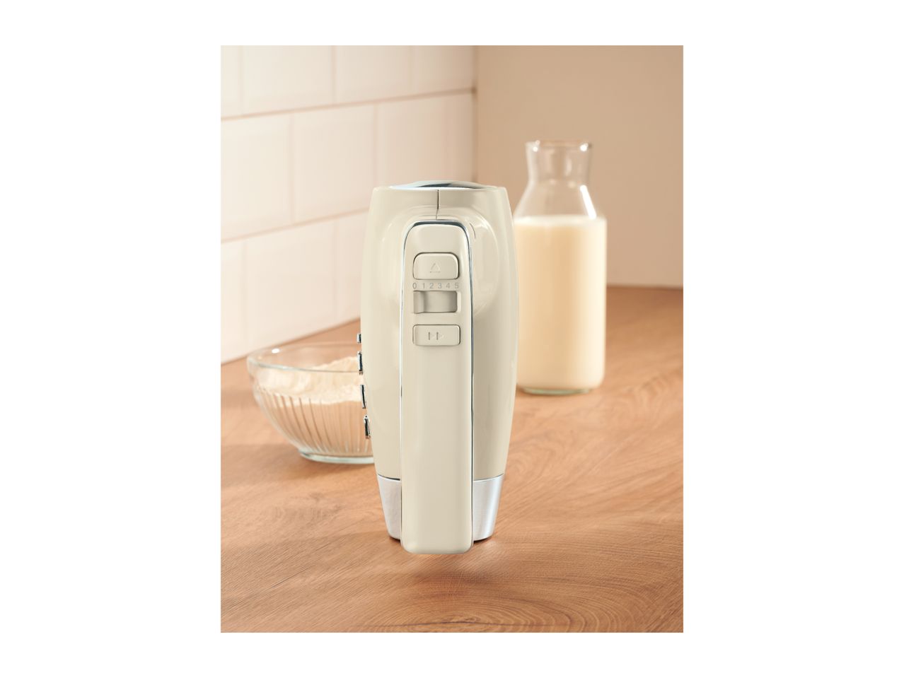 Cream-colored hand mixer standing upright on a wooden countertop next to baking ingredients.