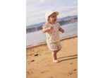 Happy baby in a sun hat and floral outfit walking on a sandy beach.