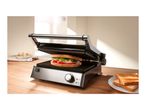 Open grill with a sandwich inside, next to a plate of toast on a wooden table.
