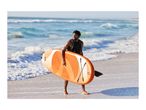 Man in a life vest carrying an orange paddleboard on a sandy beach with ocean waves.