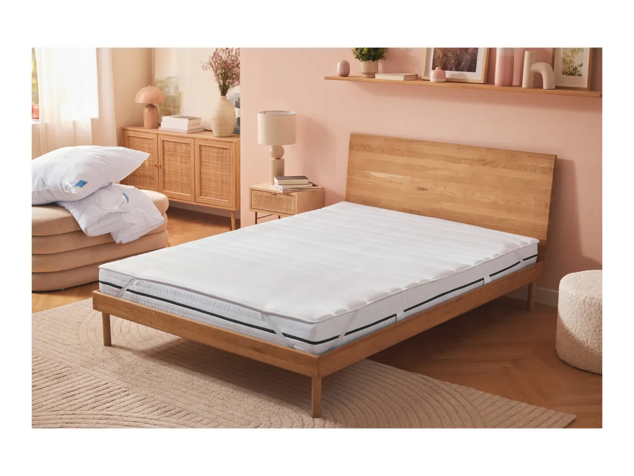 Bedroom with a wooden bed frame, white mattress, and pillows on a beige ottoman, featuring a pink wall and wooden furniture.