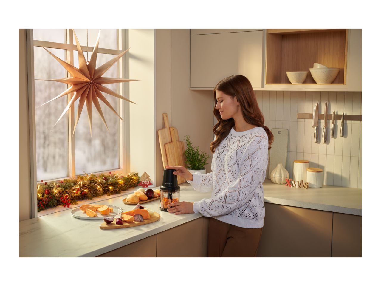 A woman using a compact food chopper in a cozy, festively decorated kitchen.